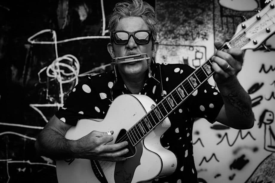 G Love with a harmonica in his mouth and guitar in hand.